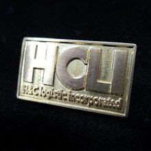 「真鍮ピンバッジセット」のHCLIロゴ。 (C)2012 高橋慶太郎・小学館／ヨルムンガンド製作委員会