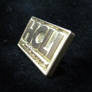 「真鍮ピンバッジセット」のHCLIロゴ。 (C)2012 高橋慶太郎・小学館／ヨルムンガンド製作委員会