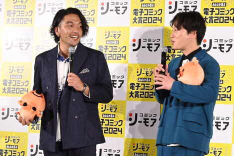 コミックシーモアのマスコットキャラクター・ヨムビーのぬいぐるみを抱える見取り図。