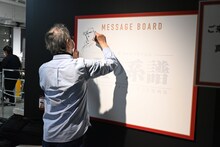 「Kの系譜」の内覧会にて、メッセージボードにイラストとサインを描き込む真船一雄。
