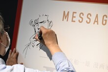 「Kの系譜」の内覧会にて、メッセージボードにイラストとサインを描き込む真船一雄。