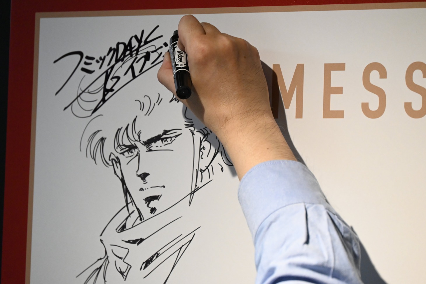 「Kの系譜」の内覧会にて、メッセージボードにイラストとサインを描き込む真船一雄。
