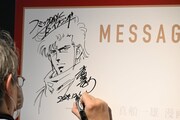 「Kの系譜」の内覧会にて、メッセージボードにイラストとサインを描き込む真船一雄。
