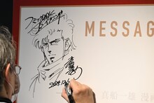 「Kの系譜」の内覧会にて、メッセージボードにイラストとサインを描き込む真船一雄。