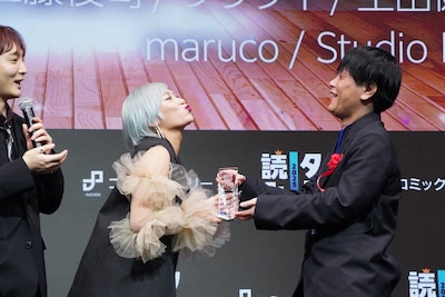 「俺だけ最強超越者～全世界のチート師匠に認められた～」の作者である江藤俊司（右）にキスを贈ろうとする信子（左）