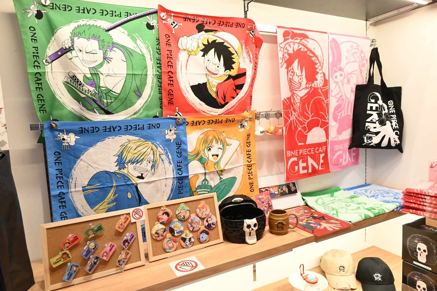 4月1日より販売される「ONE PIECE CAFE GENE」の新グッズ、再入荷グッズ。右側にかけられているフェイスタオル、平置きされているメタリッククリアファイル、「ブルックヘッドボウル」、「樽マグ」は新発売。左にかかっている和風バンダナは再販売。
