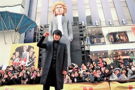 巨大龍彦像の前でポーズを取る綾野剛。