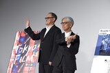 ムービーカメラに向かって手を振るスティーブン・ノムラ・シブル（左）と、坂本龍一（右）。