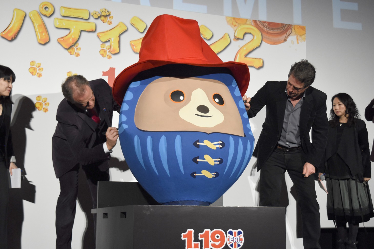 パディントンだるまにサインを入れるヒュー・ボネヴィル（左）と、ヒュー・グラント（右）。