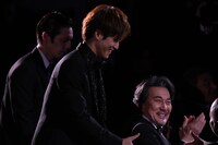 名前を呼ばれて席を立つ松坂桃李（左）と、笑顔で祝福する役所広司（右）。