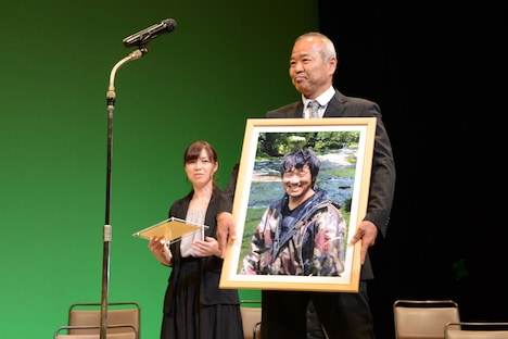 舟山弘一の写真とともに登壇する辻井啓司（手前）と、代理登壇した舟山の夫人（奥）。