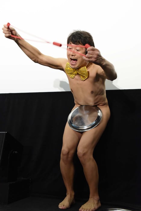 大橋彰（アキラ100%）がお盆を落とさないように筋トレ用のゴムを引っ張る様子。