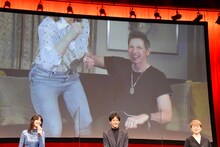 「みんなが立ってるから、私たちも立ったほうがいいかしら？」と腰を浮かせるミラ・ジョヴォヴィッチ（スクリーン左）と、それを止めるポール・W・S・アンダーソン（スクリーン右）。