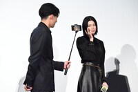 水上恒司（手前）が手にしたカメラにポーズを決める三吉彩花（奥）