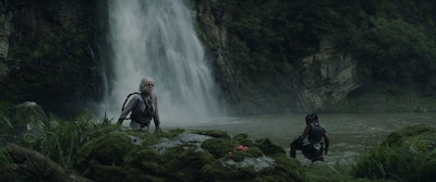 映画「プレデター：バッドランド」から、滝壺のほとりに佇むデク（ディミトリウス・コロアマタンギ / 右）とティア（エル・ファニング / 左）