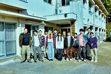 映画「四月の余白」場面写真。全寮制更生施設みらいの里の前で、一ノ瀬ワタル演じる寮長・西健吾と職員、寮生たちが集合した様子