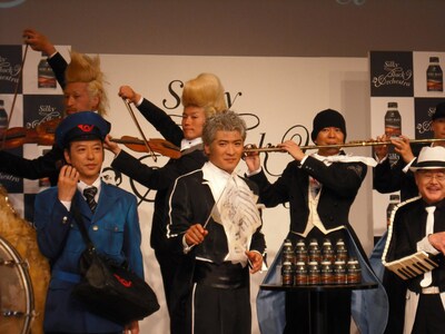 タクトを振りかざす姿も勇ましい吉川晃司（写真中央）や、なぜか郵便局員姿ルックの板尾創路（写真左端手前）。どのような演奏が繰り広げられているのかはCMでご確認を。