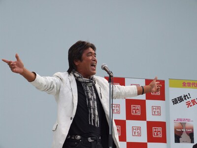 白いジャケット、黒いシャツ、褐色の肌。今年還暦とは思えない元気な姿で「ニッポンに元気を！」とエールを送るしげる。