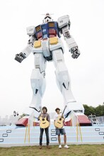 潮風公園に降り立った実物大ガンダムの前でアコギを掲げるDEPAPEPEの2人。徳岡慶也（写真右）は、巨大なガンダムを背後に心なしか不安そうな表情を浮かべている。