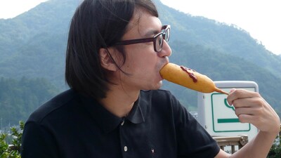 写真は本編映像より。アメリカンドッグにパクつくフルカワトオル35歳。