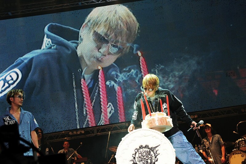 ドラム型バースデーケーキにともされたキャンドルの火を吹き消す川西幸一50歳と、それをあたたかく見守るユニコーンのメンバー。キャンドルは全部で5本、1本で10歳換算だ。