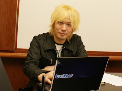 Twitterステッカーが貼られたPCを前にはにかむ津田大介。