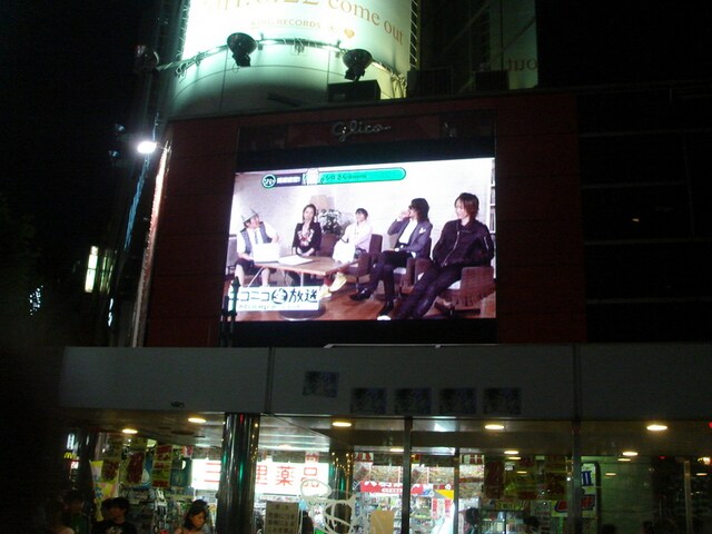 写真は渋谷の街頭ビジョンで行われたサプライズ生中継の模様。