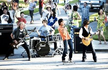 広大な住宅街セットの中で熱演するB'z。バックを務めるのは彼らのライブではおなじみのシェーン・ガラス（Dr）とバリー・スパークス（B）の2人。