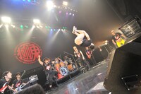 團長の無茶ぶりに応え、打点の高いバク宙を決める石鹸。ステージの左右ではメンバーの港カヲル（写真右端）とバイト君（写真左端）がまじまじとみつめている。