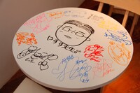 メンバー直筆のイラスト入りテーブル。中央には片山陽加画伯による「やすす先生（秋元康）」の似顔絵が描かれている。
