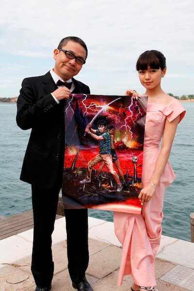 ベネチアの地で「地獄でなぜ悪い」のジャケットにも使用されたポスターとともに写真に収まる園子温と二階堂ふみ（左から）。