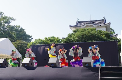 金の鯱がきらめく名古屋城をバックに歌うチームしゃちほこ。