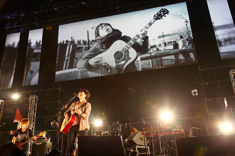 過去の路上ライブ映像をバックにメジャーデビュー曲「素晴らしき日常」を熱唱する高橋優。