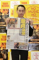 自身の童貞偽装を報じる新聞の号外風フライヤーとミニアルバム「あの娘のメルアド予想する」を手に写真に収まる柴田隆浩。