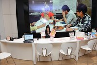 受付嬢になりきる伊藤千由李。背後の巨大ディスプレイにはチームしゃちほこ「首都移転計画」のPVが。