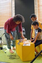 子供たちのオリジナル楽器にアドバイスするU-zhaan（左）。