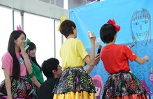 高城れにの似顔絵を描く百田夏菜子（右）と、それを見つめるメンバー。