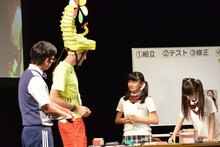 流暢な英語でジョセフ・テイムと会話する日高麻鈴と、その横で黙々と作業を続ける吉田爽葉香。