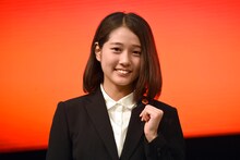 リクルートスーツの衣装にあしらわれた“つばきファクトリー社”のバッジを見せる谷本安美。