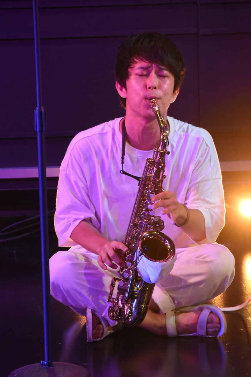 座りながらサックスを吹く塩チョ（Sax, Fl / 西村ヒロチョ）。