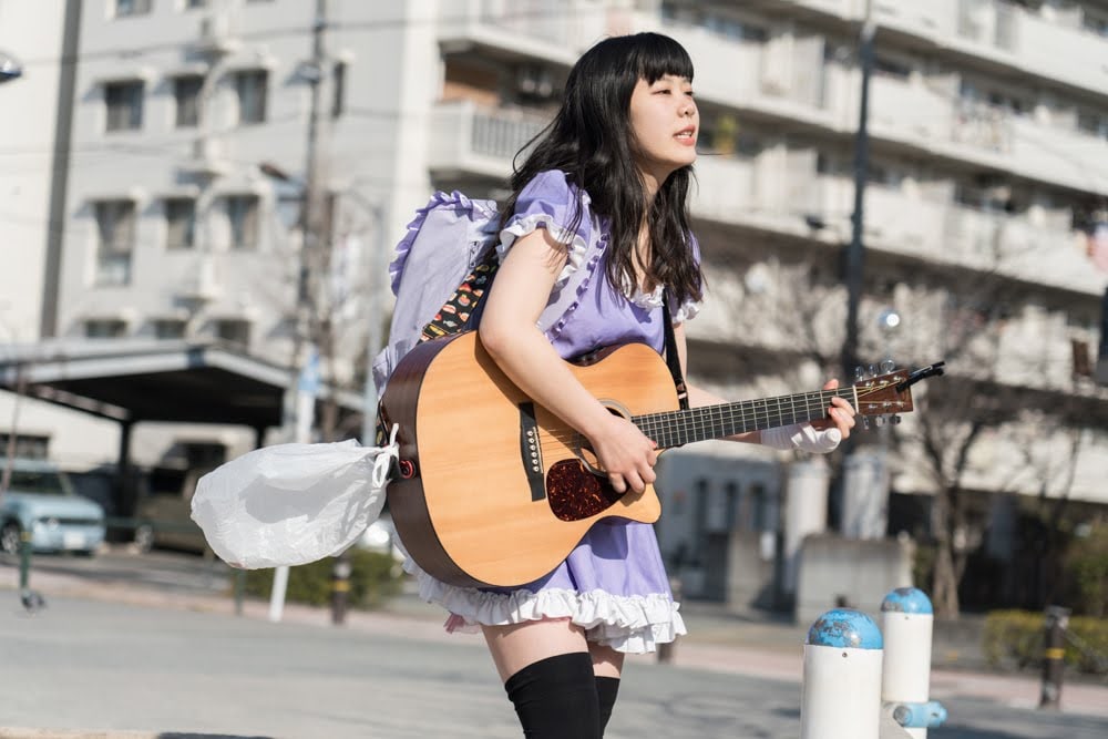 街中をギターを弾き語りながら歩く眉村ちあき。