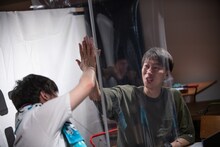 パーテーション越しにハイタッチするグランジ遠山（左）とはんにゃ金田（右）。