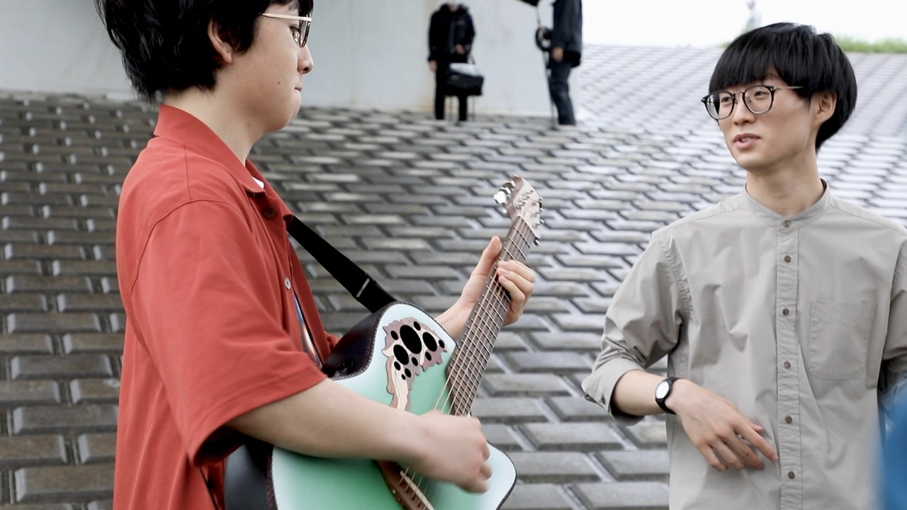 河川敷で歌う崎山蒼志（左）と、それを見守る山﨑すがら（右）。