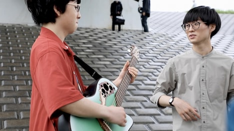 河川敷で歌う崎山蒼志（左）と、それを見守る山﨑すがら（右）。