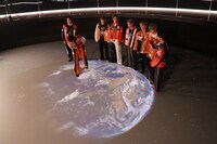 床面スクリーンに映し出される地球の上に立つメンバー。