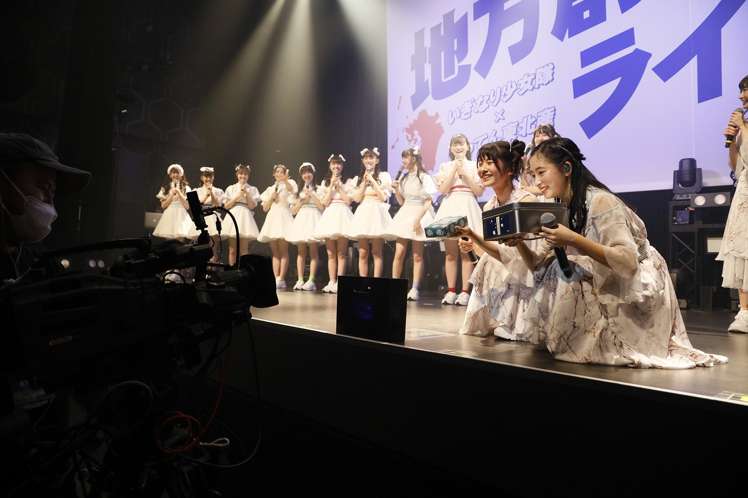 もち吉のおせんべいと博多あまびをカメラに向けるばってん少女隊の蒼井りるあ、柳美舞。