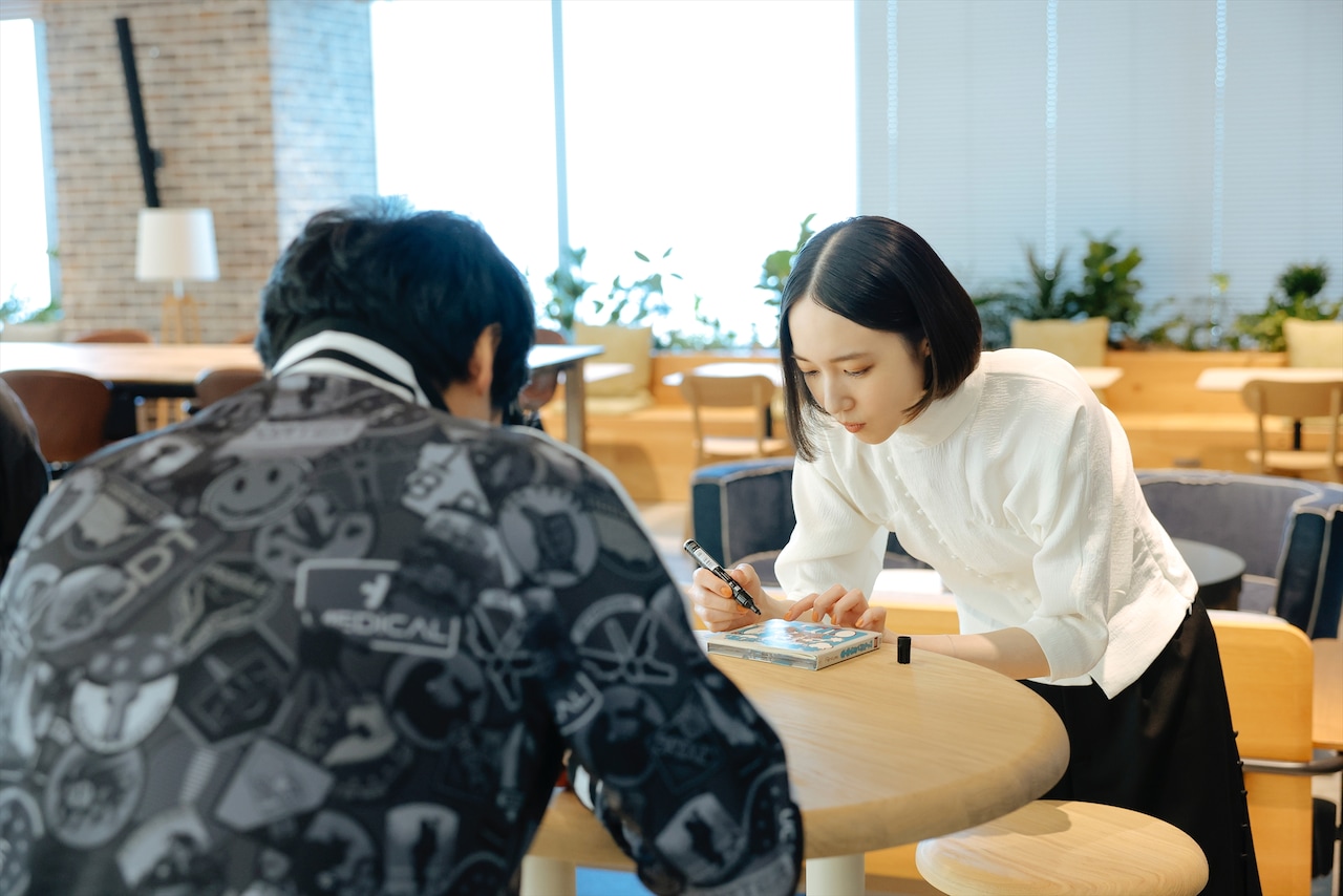 のっちさん私物のBBポッド アクリルキーホルダーにサインする小島秀夫さんと、小島さん私物のPerfume「未来のミュージアム」シングルジャケットにサインするのっちさん。