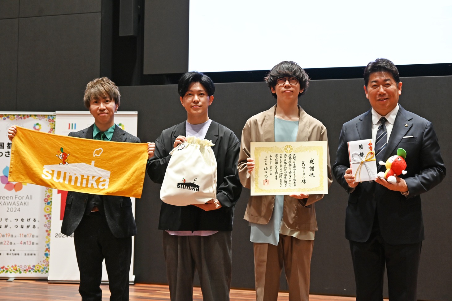 左からコラボグッズを持った荒井智之と小川貴之、感謝状を持った片岡健太、目録を持った福田紀彦氏。