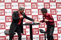 テーブルに固定されていた広島グルメの皿を引き剥がして報道陣に見せる吉川晃司（左）。