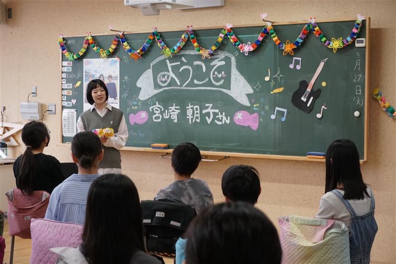 児童との交流会で「ようこそ宮崎朝子さん」と装飾された黒板を前に立つ、宮崎朝子（SHISHAMO / G, Vo）。